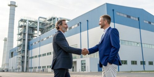 Two business leaders shaking hands in front of a modern factory.