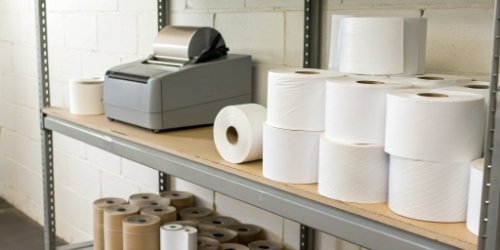 Label Inventory Cost Savings A shelf overflowing with various label rolls on one side, and a neat stack of blank label rolls next to a scale printer on the other.