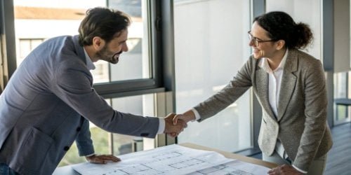 Two business professionals shaking hands over a table with technical diagrams.