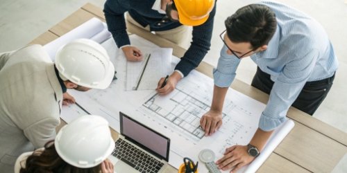 A team of engineers and managers collaborating around a table with blueprints and a laptop.